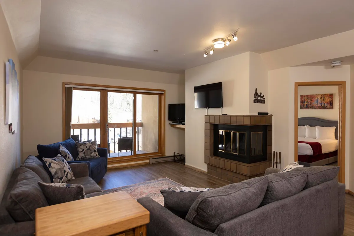 A living space with a cozy fireplace, soft sofa, large window offering mountain views, and a flat-screen TV, perfect for unwinding after a day on the slopes. The warm lighting and arrangement create a welcoming atmosphere for group relaxation and entertainment.