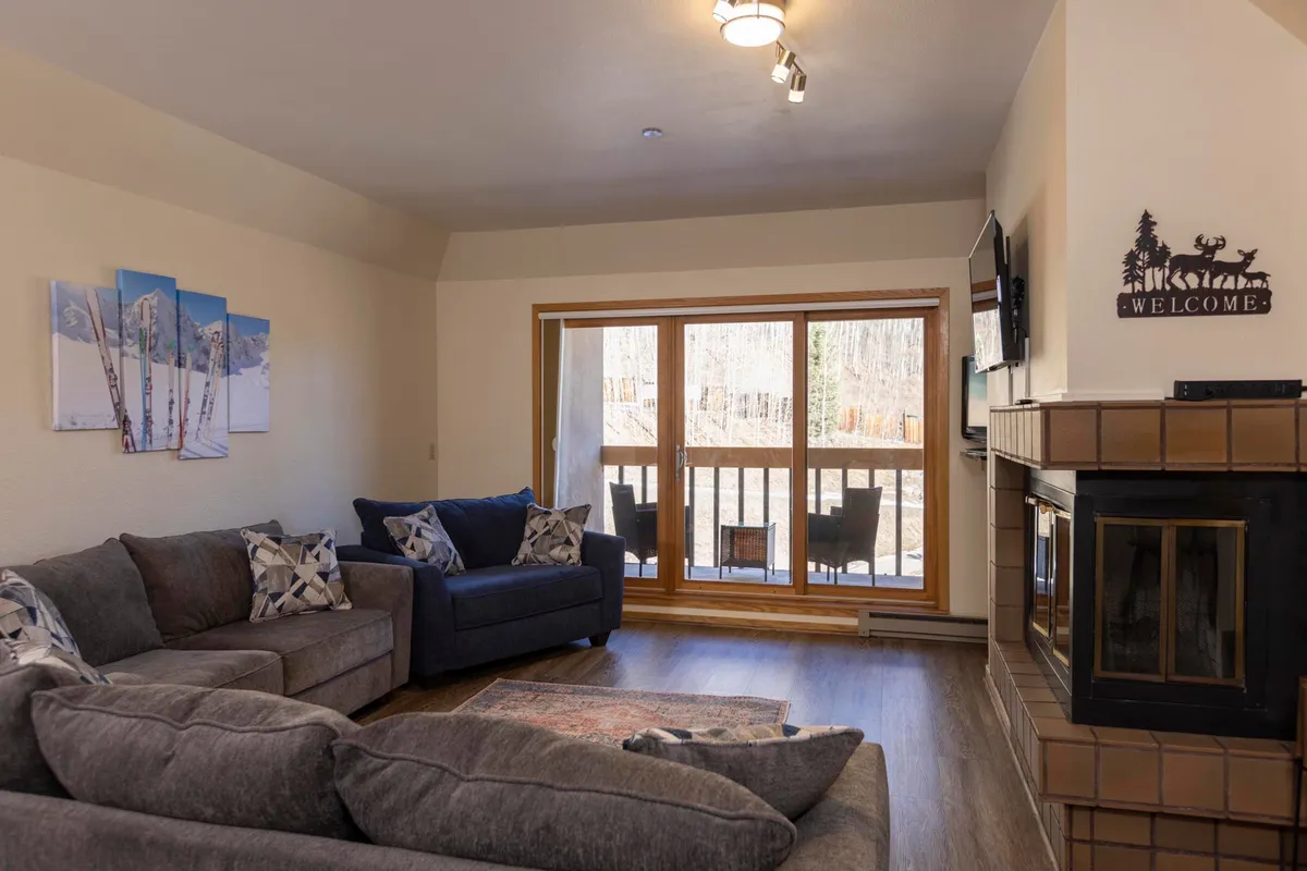 A welcoming living area with comfortable sofas arranged around a stone fireplace, large windows framing panoramic mountain views, and tasteful artwork on the wall. The space is bathed in natural light, inviting relaxation and socializing.