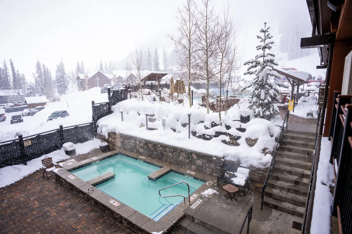 A heated pool and hot tub are surrounded by snow-covered trees, with steam rising from the water and people relaxing in the warm water. The scene conveys a blend of winter scenery and resort comfort, with steps leading down to the pool area, emphasizing both accessibility and tranquility.