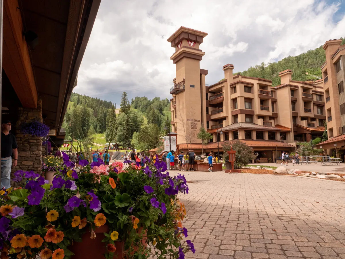 This lively exterior image features the tall Village Center building with its recognizable clock tower, colorful flower pots, and people enjoying the outdoor communal space. The scene is vibrant and welcoming, emphasizing the social and lively atmosphere of the village center.