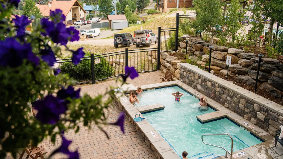 An inviting outdoor rooftop hot tub, surrounded by people relaxing and socializing, with expansive mountain views and bright landscaping featuring colorful flowers. The setting glows with leisure and warmth, even in the brisk mountain air.