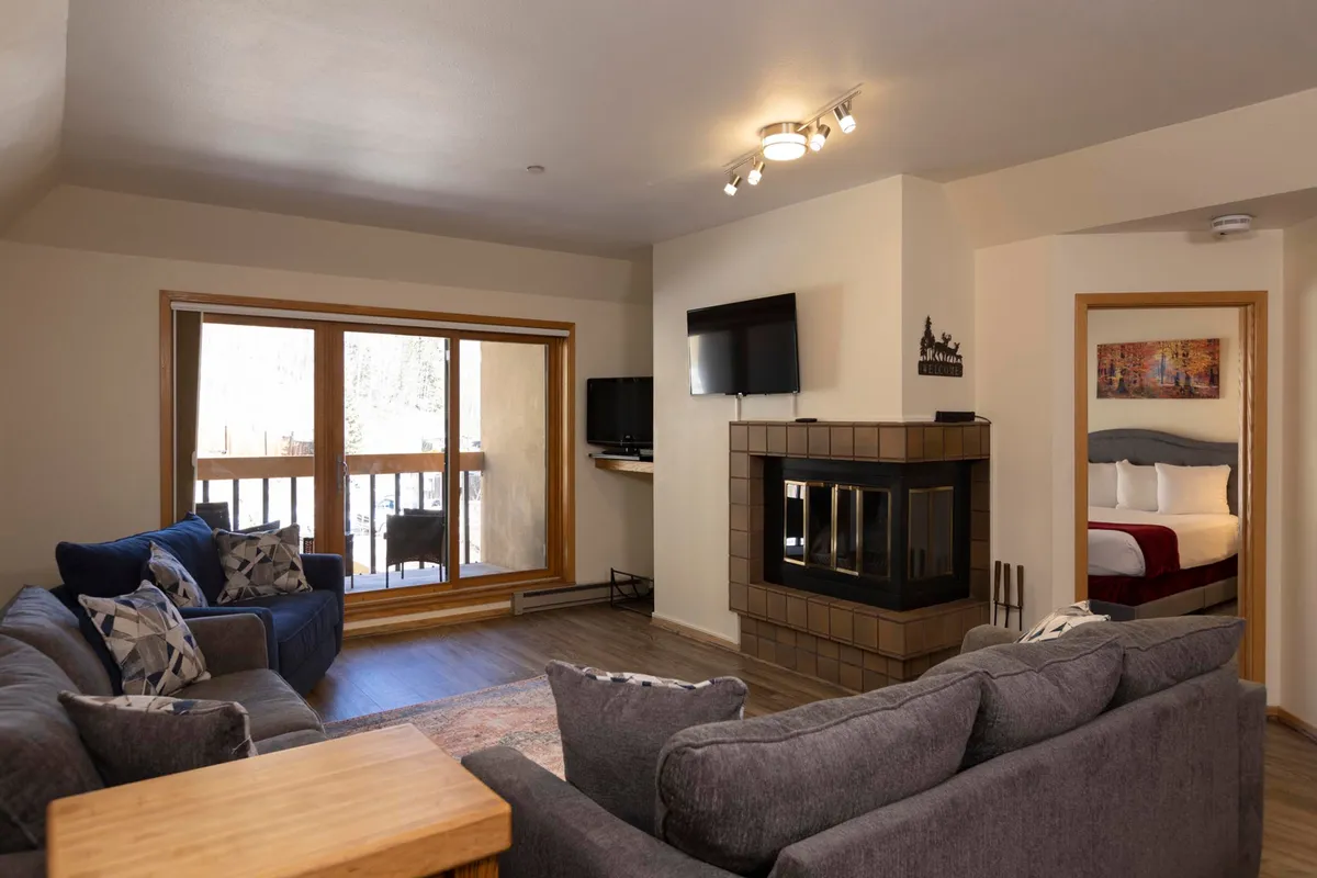 The image showcases a cozy living room with plush seating arranged around a glowing fireplace, large windows framing breathtaking mountain views, and a television for entertainment. The space feels inviting, with ample natural light, soft blankets, and a welcoming atmosphere perfect for relaxing after a day on the slopes.