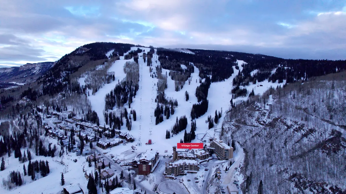 A panoramic view of snow-covered ski slopes with a prominently placed 'Village Center' sign, surrounded by frosted trees and a dramatic mountain range in the distance. The image exudes the energy of a busy winter day at the heart of the resort community.