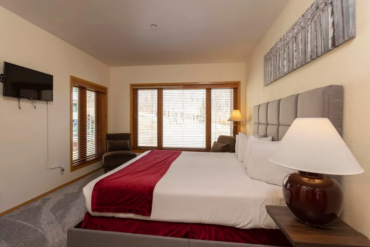 A spacious bedroom with a king-sized bed dressed in red and white linens, large windows with wooden blinds, an armchair in the corner, and a wall-mounted TV. Soft lighting and subtle decor create an inviting and restful environment ideal for couples or small groups.