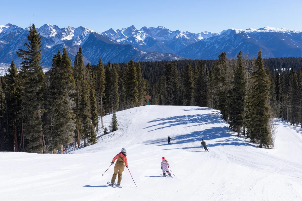 A dynamic winter scene featuring skiers descending a snow-covered slope, with majestic mountains and tall pine trees in the background. The snow appears freshly groomed, and the scene is vibrant with families and friends enjoying an exhilarating day on the mountain.