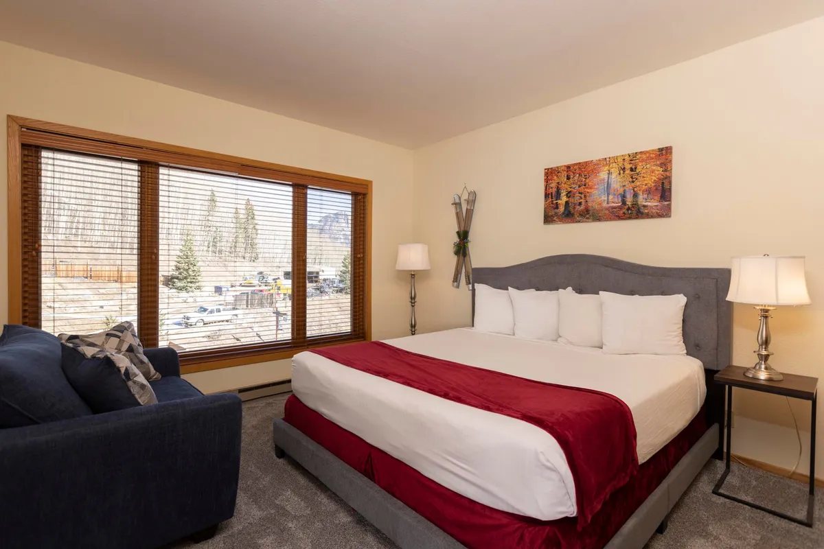 A tranquil bedroom with a large window offering breathtaking mountain views. The space features a comfortable bed with a striking red accent, a cozy armchair by the window, and soft lighting that creates a serene atmosphere. The overall mood is peaceful, perfect for unwinding after a day of adventure.