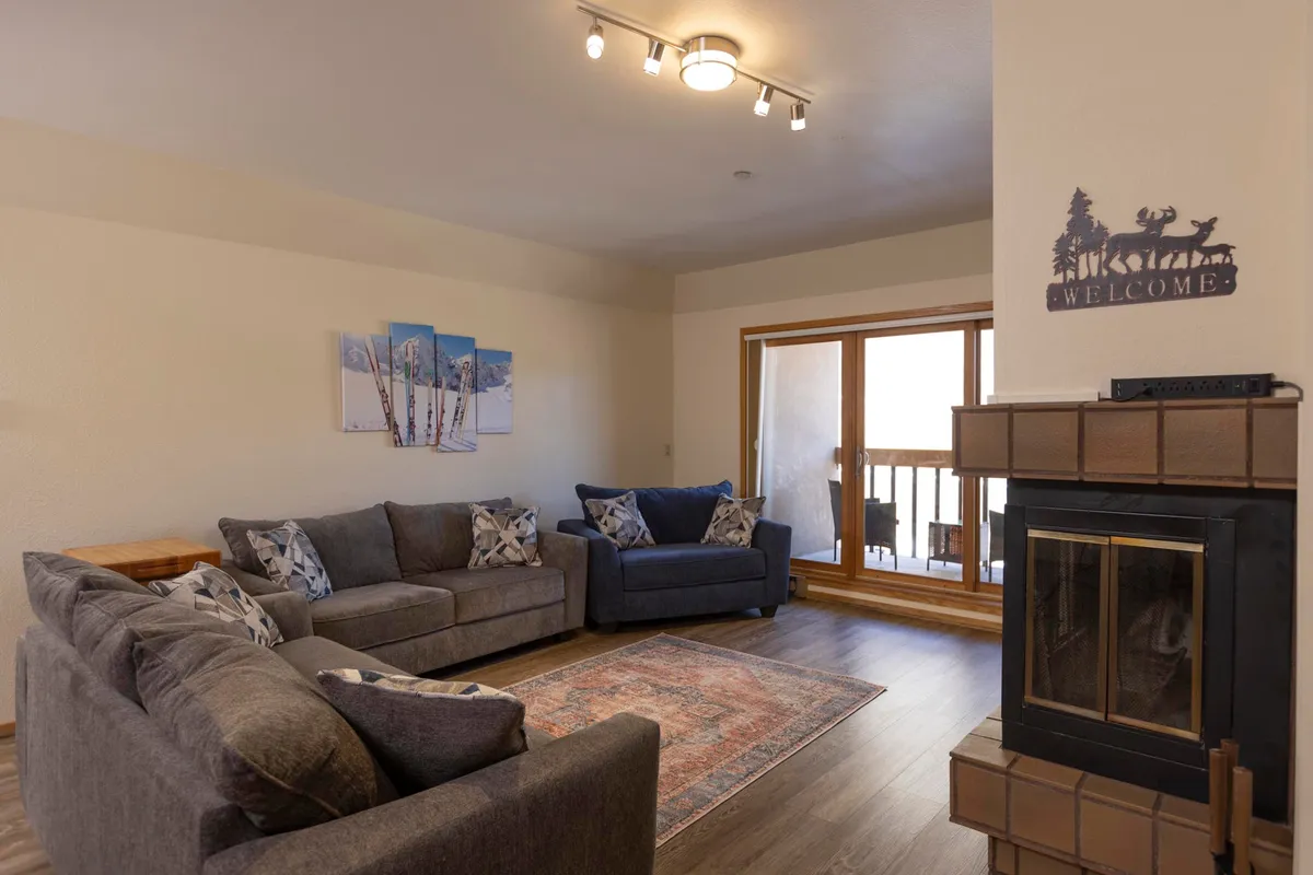 A comfortable, inviting living room featuring sofas set around a fireplace, a large scenic window that brings the outdoors in, and tasteful wall art. The atmosphere is designed for socializing and enjoying the mountain surroundings.