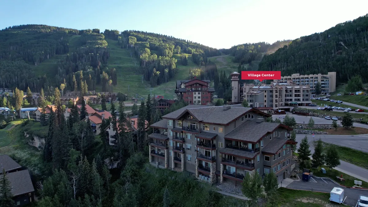 An exterior shot showing the surrounding mountains, multiple buildings, and accessible parking area within the mountain village. The scene combines natural beauty with direct access to recreational activities, giving a sense of both adventure and practicality.