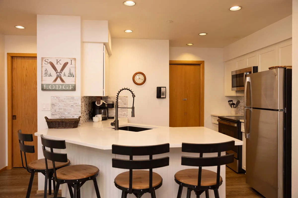 A warm, well-lit kitchen with a central island, bar stools, visible kitchen appliances, and a 'Ski Lodge' sign. The setup is ideal for casual dining, entertaining, and creating culinary memories after a day outdoors. It radiates a homey, inviting ambiance.